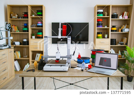 A 3D printer is actively creating a red object on a desk with spools of filament and a laptop displaying a 3D model A 3D printer is actively creating a red object on a desk with spools of filament and a laptop displaying a 3D model 137771595