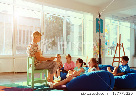 Teacher reading a story to attentive children during cozy classroom story time in a bright kindergarten Teacher reading a story to attentive children during cozy classroom story time in a bright kindergarten 137773220