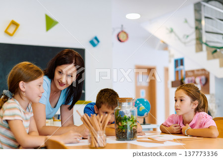 Teacher guiding elementary students as they observe a classroom terrarium and work on a science activity together Teacher guiding elementary students as they observe a classroom terrarium and work on a science activity together 137773336