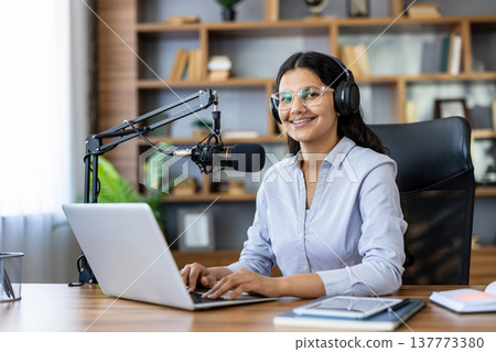 Young smiling woman wearing headphones and glasses broadcasting a podcast from a home studio, sitting at a wooden desk with a laptop and professional microphone Young smiling woman wearing headphones and glasses broadcasting a podcast from a home studio, sitting at a wooden desk with a laptop and professional microphone 137773380
