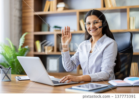 Young indian woman smiling and waving at camera while working on her laptop with a headset on during a video conference, representing remote work, online education, and virtual communication Young indian woman smiling and waving at camera while working on her laptop with a headset on during a video conference, representing remote work, online education, and virtual communication 137773391