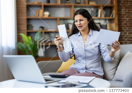 Young woman at home stressed over financial bills and receipts, sitting on sofa with laptop and paperwork, worried about debt, budgeting and expenses while working remotely Young woman at home stressed over financial bills and receipts, sitting on sofa with laptop and paperwork, worried about debt, budgeting and expenses while working remotely 137773403