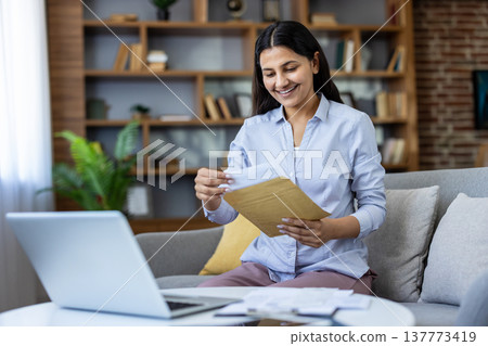 Young woman smiling while opening a brown envelope with a letter, sitting on a sofa in a home office environment with a laptop and paperwork on a coffee table 137773419