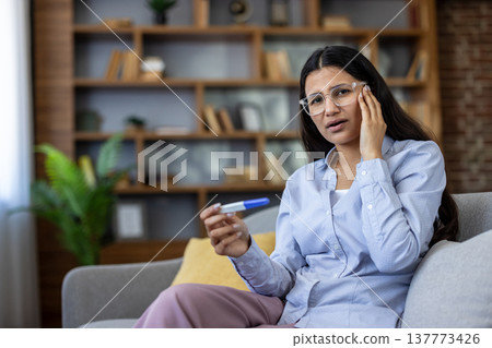 Young woman sitting on a sofa at home, holding a pregnancy test and showing a surprised and worried expression, reflecting on unexpected life changes and future plans Young woman sitting on a sofa at home, holding a pregnancy test and showing a surprised and worried expression, reflecting on unexpected life changes and future plans 137773426
