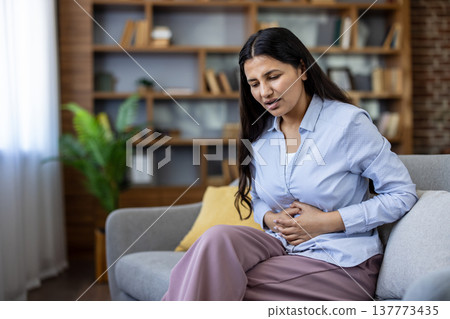 Young indian woman sitting on a sofa and holding her stomach, experiencing severe abdominal pain, indigestion, or menstrual cramps, highlighting health issues and discomfort Young indian woman sitting on a sofa and holding her stomach, experiencing severe abdominal pain, indigestion, or menstrual cramps, highlighting health issues and discomfort 137773435