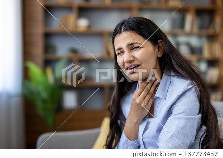 Young woman at home clutching her throat and neck in discomfort, showing symptoms of sore throat or infection, unwell and needing medical care while sitting on a couch 137773437