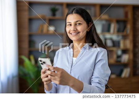 Young indian woman smiling and holding a smartphone, actively engaging with technology, communication, and digital connectivity in a modern office or home setting 137773475