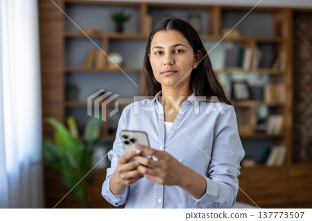 Young woman standing in a home office, holding a smartphone, representing modern communication, online work, and digital technology in a remote workspace Young woman standing in a home office, holding a smartphone, representing modern communication, online work, and digital technology in a remote workspace 137773507