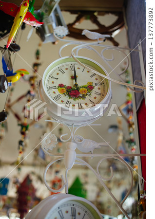 A vertical shot of a white decorative double-sided hanging clock featuring a floral pattern on the dial and ornate wrought-iron style mounting, displayed in a gift shop. A vertical shot of a white decorative double-sided hanging clock featuring a floral pattern on the dial and ornate wrought-iron style mounting, displayed in a gift shop. 137773892