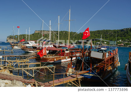 A wide horizontal shot of several wooden excursion boats with Turkish flags docked at a wooden pier in a turquoise sea bay against green mountains and a clear blue sky. A wide horizontal shot of several wooden excursion boats with Turkish flags docked at a wooden pier in a turquoise sea bay against green mountains and a clear blue sky. 137773896