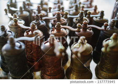 A close-up horizontal shot of multiple vintage-style metal coffee or pepper mills with ornate engravings, arranged in rows on a shop shelf. A close-up horizontal shot of multiple vintage-style metal coffee or pepper mills with ornate engravings, arranged in rows on a shop shelf. 137773945
