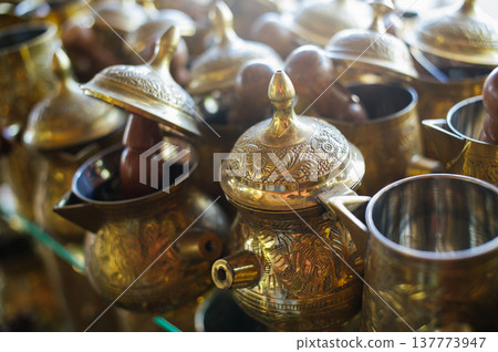A close-up horizontal shot of handcrafted brass coffee pots (cezve) with intricate floral engravings and pointed lids, displayed on a shop shelf. A close-up horizontal shot of handcrafted brass coffee pots (cezve) with intricate floral engravings and pointed lids, displayed on a shop shelf. 137773947