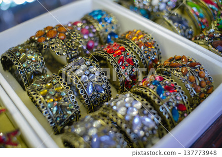 A close-up horizontal shot of multiple wide, open-work metal bracelets decorated with vibrant colorful crystals and stones, neatly arranged in a white display box. A close-up horizontal shot of multiple wide, open-work metal bracelets decorated with vibrant colorful crystals and stones, neatly arranged in a white display box. 137773948