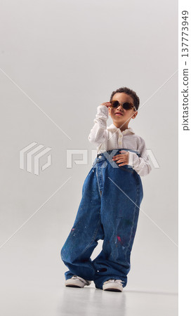 Little boy wearing oversized denim jeans and sunglasses posing confidently in studio. Little boy wearing oversized denim jeans and sunglasses posing confidently in studio. 137773949