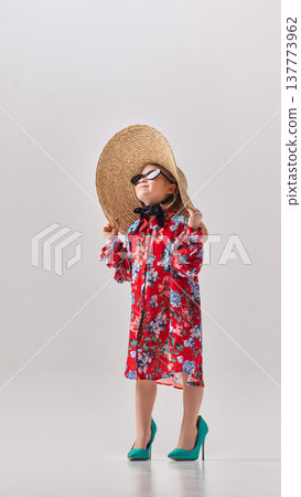 Little girl wearing oversized floral dress straw hat and high heels standing in studio looking upward Little girl wearing oversized floral dress straw hat and high heels standing in studio looking upward 137773962