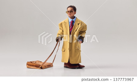 Little boy wearing oversized coat and sunglasses dragging large leather briefcase across studio floor. Little boy wearing oversized coat and sunglasses dragging large leather briefcase across studio floor. 137773967