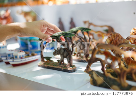 A close-up horizontal shot of a person's hand reaching for a small, detailed elephant figurine placed under a stylized wooden tree canopy, with other animal statues blurred in the background. A close-up horizontal shot of a person's hand reaching for a small, detailed elephant figurine placed under a stylized wooden tree canopy, with other animal statues blurred in the background. 137773976
