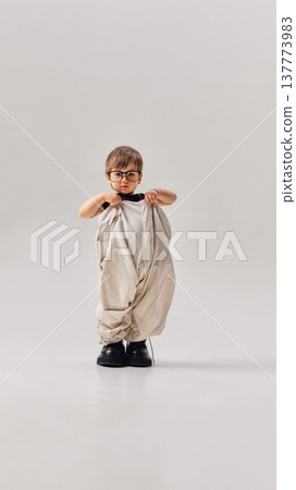 Little boy wearing oversized adult trousers and large shoes standing in studio. Little boy wearing oversized adult trousers and large shoes standing in studio. 137773983