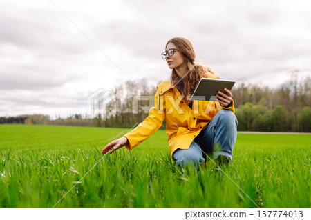 Young farmer woman in a green field, intently using a tablet. She wears a yellow jacket and glasses Young farmer woman in a green field, intently using a tablet. She wears a yellow jacket and glasses 137774013