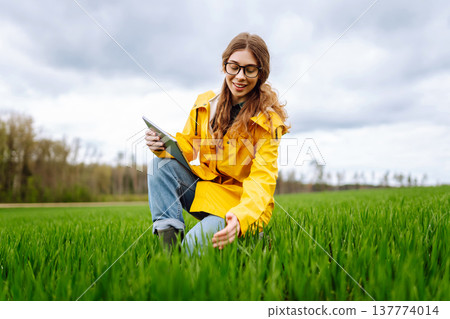 Young farmer woman in a green field, intently using a tablet. She wears a yellow jacket and glasses Young farmer woman in a green field, intently using a tablet. She wears a yellow jacket and glasses 137774014