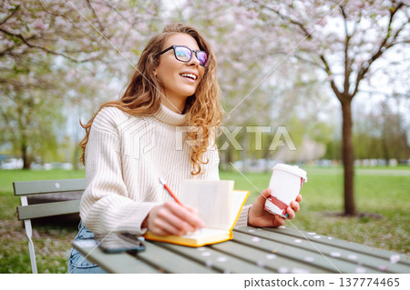 Cute woman in glasses writes in notepad, sits with laptop enjoying sunny spring day. Relax concept. 137774465