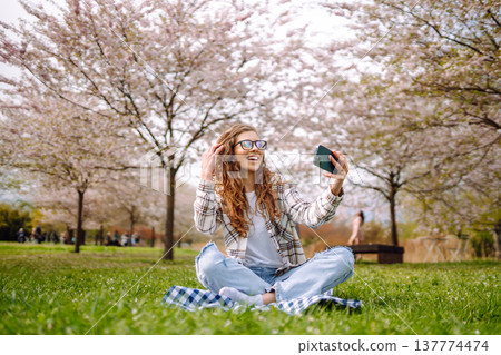 Young woman takes selfie against background of flowering trees in park. Concept of relaxation, fun. Young woman takes selfie against background of flowering trees in park. Concept of relaxation, fun. 137774474