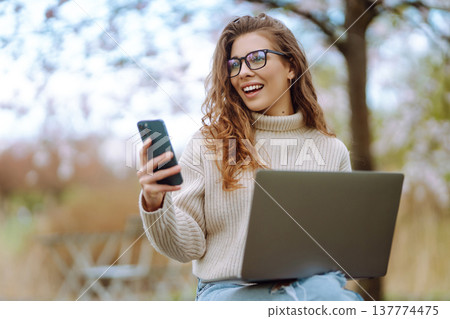 Cheerful woman on phone takes selfie while sitting at table with laptop in spring park. Lifestyle. Cheerful woman on phone takes selfie while sitting at table with laptop in spring park. Lifestyle. 137774475