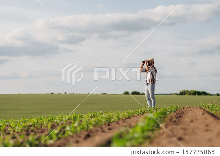 Black binoculars is in hands. Looking far away. Man farmer is on the agricultural field Black binoculars is in hands. Looking far away. Man farmer is on the agricultural field 137775063
