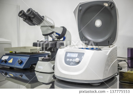A close-up of a modern laboratory workstation featuring an open benchtop centrifuge, a binocular microscope, and a laboratory stirrer on a clean counter A close-up of a modern laboratory workstation featuring an open benchtop centrifuge, a binocular microscope, and a laboratory stirrer on a clean counter 137775553