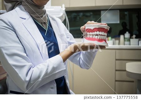 A female dentist wearing a hijab and a white lab coat holds a dental model of human teeth and gums in a clinic. She is demonstrating oral hygiene or dental health education A female dentist wearing a hijab and a white lab coat holds a dental model of human teeth and gums in a clinic. She is demonstrating oral hygiene or dental health education 137775558