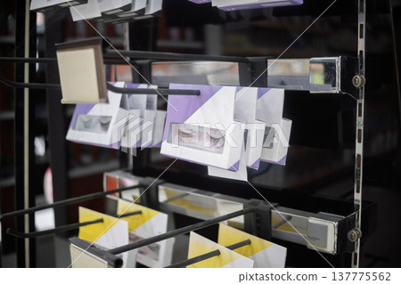 Close-up of packaged false eyelashes hanging on metal hooks in a beauty store aisle, featuring various styles in purple and yellow boxes 137775562