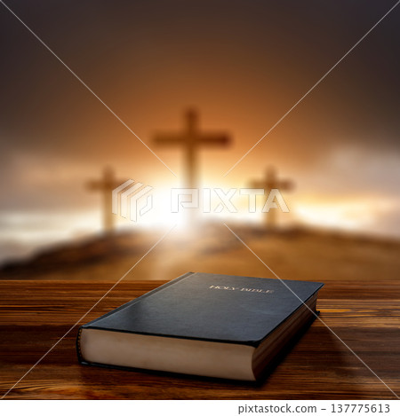 Closed Holy Bible on a wooden surface with a blurred background of three crosses on a hill during a golden sunset, symbolizing faith, sacrifice, and Christian devotion 137775613