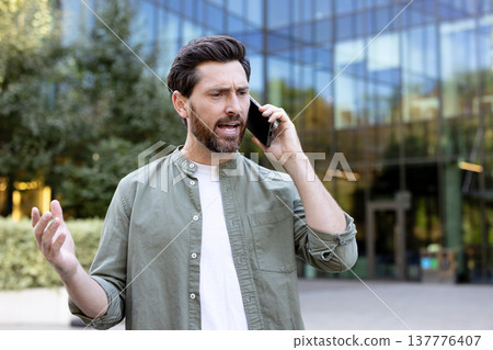 Man feeling frustration and anger during an unpleasant phone conversation, expressing disagreement and stress while communicating on a smartphone outdoors near a modern office building 137776407
