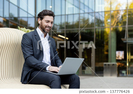 Smiling businessman in a suit and wireless earbuds types on a laptop while seated on a modern bench in a corporate park, illustrating mobile productivity and business connectivity 137776426