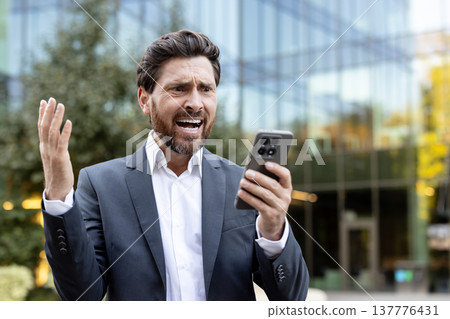 Businessman looking angry and stressed, shouting at his mobile phone while standing outside a modern office building, reacting to negative information or a failing business deal Businessman looking angry and stressed, shouting at his mobile phone while standing outside a modern office building, reacting to negative information or a failing business deal 137776431