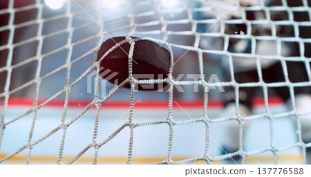 Close-up action shot of an ice hockey puck hitting the goal net during a scoring moment. Dynamic sports scene with ice spray and a blurred hockey player in the background capturing the intensity of Close-up action shot of an ice hockey puck hitting the goal net during a scoring moment. Dynamic sports scene with ice spray and a blurred hockey player in the background capturing the intensity of 137776588