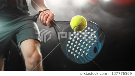Dynamic action shot of a padel player hitting a yellow ball with a racket during a match. Dramatic lighting highlights movement, speed and power, capturing the energy and intensity of modern racket 137776599