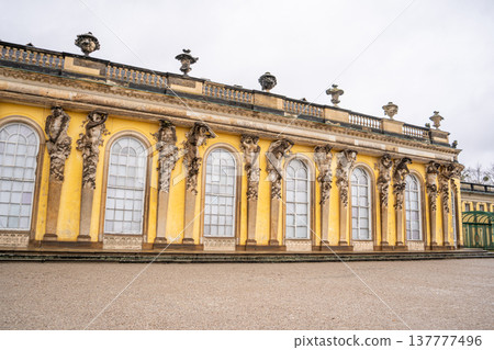 Caryatids and Atlanteans supporting the roof Sanssouci Palace in Potsdam, Germany. Old building architecture in Rococo style. Yellow facade wall and windows. Caryatids and Atlanteans supporting the roof Sanssouci Palace in Potsdam, Germany. Old building architecture in Rococo style. Yellow facade wall and windows. 137777496
