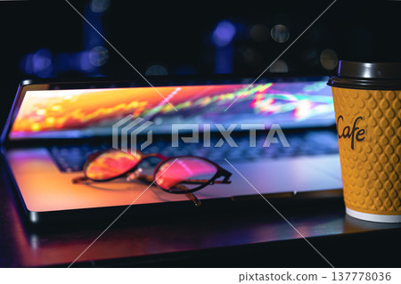 A laptop half closed in the dark with colourful glow, cup of coffee and glasses. A laptop half closed in the dark with colourful glow, cup of coffee and glasses. 137778036