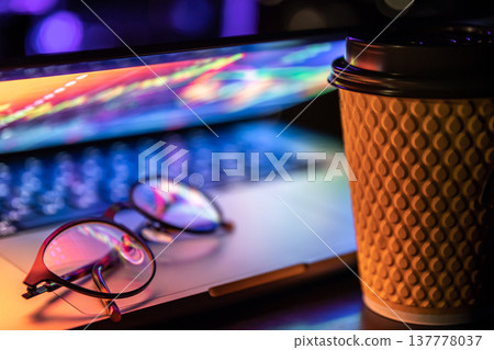 A laptop half closed in the dark with colourful glow, cup of coffee and glasses. A laptop half closed in the dark with colourful glow, cup of coffee and glasses. 137778037