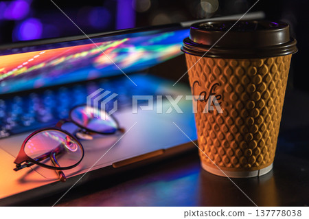 A laptop half closed in the dark with colourful glow, cup of coffee and glasses. A laptop half closed in the dark with colourful glow, cup of coffee and glasses. 137778038