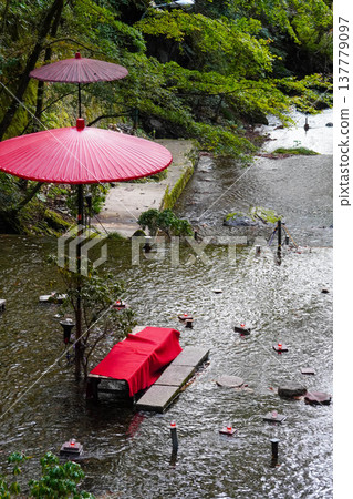 A bench covered with a red felt mat and a red Japanese umbrella are set up in the middle of the Kifune River. A bench covered with a red felt mat and a red Japanese umbrella are set up in the middle of the Kifune River. 137779097