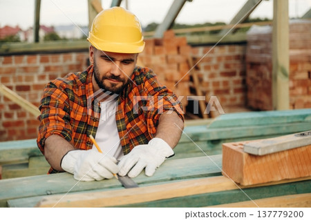 Pencil is in hand, marking place on plank. Construction worker is on the roof of a house Pencil is in hand, marking place on plank. Construction worker is on the roof of a house 137779200