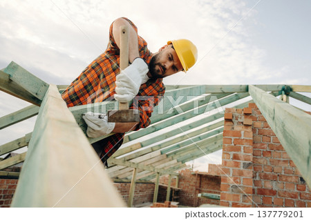 Hammer in hand, standing high up. Construction worker is on the roof of a house 137779201