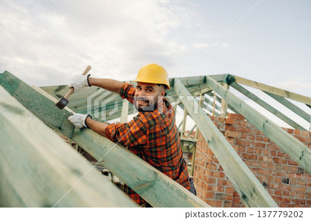 Hammer in hand, standing high up. Construction worker is on the roof of a house Hammer in hand, standing high up. Construction worker is on the roof of a house 137779202