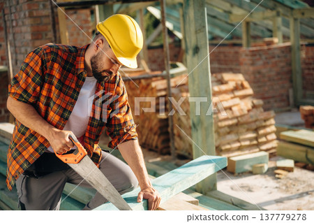 Cuts the plank with a saw. Construction worker is on the roof of a house Cuts the plank with a saw. Construction worker is on the roof of a house 137779278