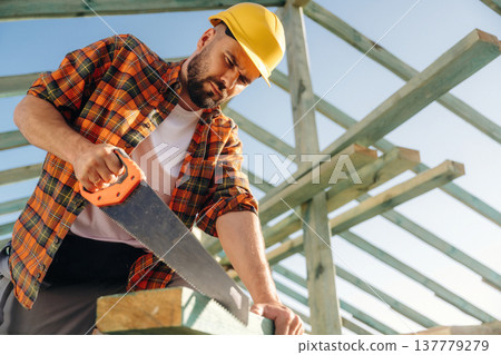 Cuts the plank with a saw. Construction worker is on the roof of a house Cuts the plank with a saw. Construction worker is on the roof of a house 137779279