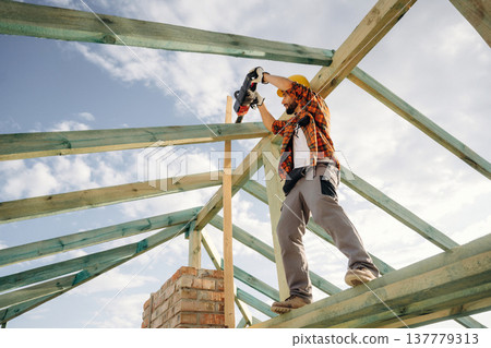 Electric chainsaw is in hands. Construction worker is on the roof of a house Electric chainsaw is in hands. Construction worker is on the roof of a house 137779313