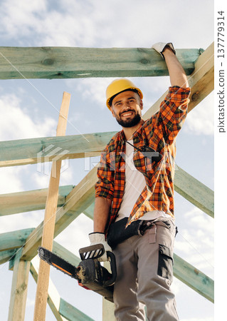 Electric chainsaw is in hands. Construction worker is on the roof of a house Electric chainsaw is in hands. Construction worker is on the roof of a house 137779314