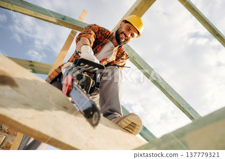 Looks down, cutting the plank with chainsaw. Construction worker is on the roof of a house Looks down, cutting the plank with chainsaw. Construction worker is on the roof of a house 137779321